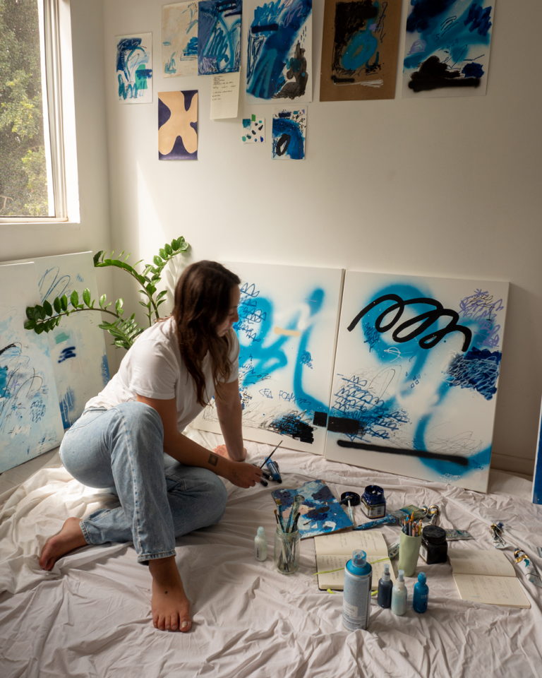 Woman sits in artist studio surrounded by paintings, paints and brushes.